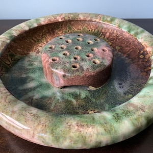 May include: A green and brown ceramic flower frog with a circular base and a raised center with holes for flower stems.