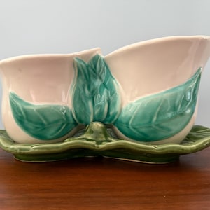 May include: A vintage ceramic planter with a double-bowl design, featuring a soft pink interior and exterior with green leaf accents. The planter sits on a matching green leaf-shaped base, perfect for displaying small plants or flowers.