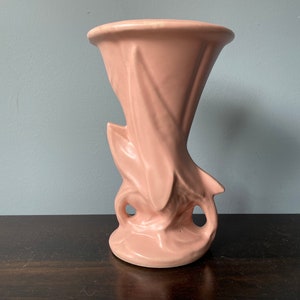 May include: A pink ceramic vase with a unique, abstract design. The vase has a wide opening and a narrow base, with a sculpted, organic shape.
