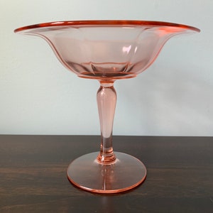 May include: A pink glass pedestal bowl with a fluted design. The bowl has a wide, shallow base and a tall, slender stem.
