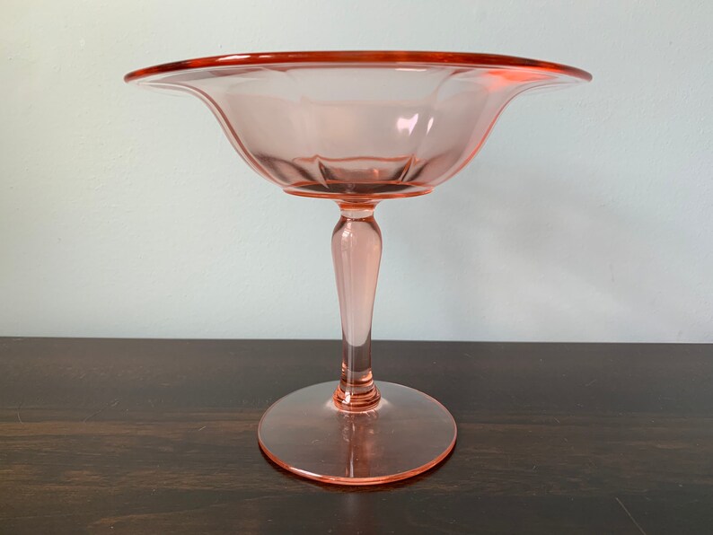 May include: A pink glass pedestal bowl with a fluted design. The bowl has a wide, shallow base and a tall, slender stem.
