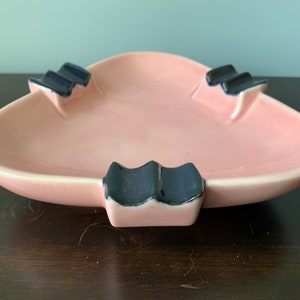 May include: A pink ceramic ashtray with three black and blue cigarette rests. The ashtray is shaped like a triangle.