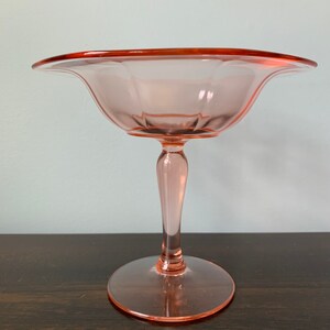 May include: A pink glass pedestal bowl with a scalloped edge and a fluted design. The bowl is on a wooden surface.