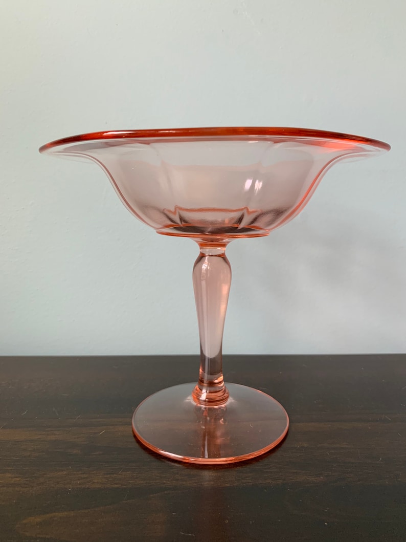 May include: A pink glass pedestal bowl with a scalloped edge and a fluted design. The bowl is on a wooden surface.