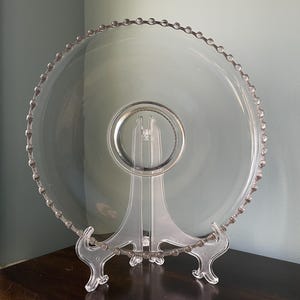 May include: A clear glass serving platter with a beaded edge and a clear glass stand. The platter is round and has a diameter of approximately 12 inches.