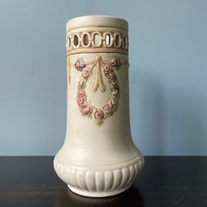 May include: A cream-colored ceramic vase with a decorative design. The vase features a detailed floral wreath with pink roses and a ribbon. The top has a series of oval cutouts. The vase has a textured surface and a bulbous base.