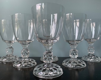 May include: Six clear glass goblets with a diamond pattern on the base and stem. The goblets are arranged in a semi-circle on a dark surface.