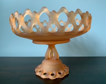 May include: A vintage peach-colored glass pedestal bowl with a delicate, openwork design. The bowl has a scalloped edge and a footed base.