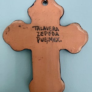 May include: A terracotta cross with blue accents and a hole at the top for hanging. The back of the cross is inscribed with "TALAVERA ZEPEDA PUE, MEX."