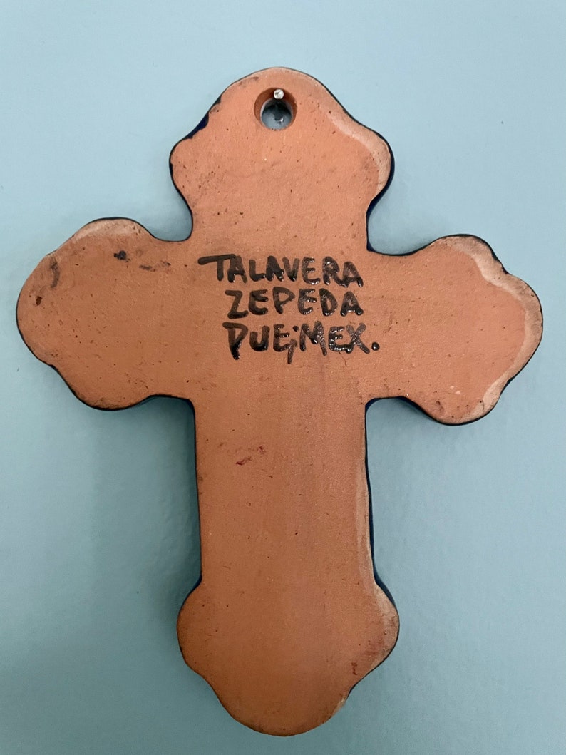 May include: A terracotta cross with blue accents and a hole at the top for hanging. The back of the cross is inscribed with "TALAVERA ZEPEDA PUE, MEX."