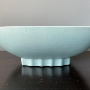 May include: A light blue ceramic bowl with a textured surface. The bowl has a wide, shallow design and sits on a scalloped base. The bowl's shape is reminiscent of a boat.