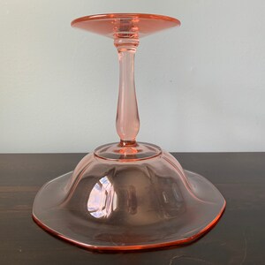 May include: A pink glass pedestal cake stand with a scalloped edge and a fluted base.
