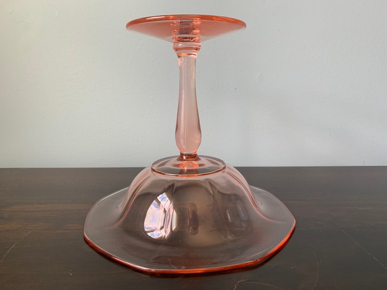May include: A pink glass pedestal cake stand with a scalloped edge and a fluted base.