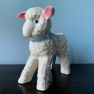 May include: White ceramic lamb planter with pink ears and a blue ribbon around its neck. The lamb has a blue eye and is standing on a dark brown surface.