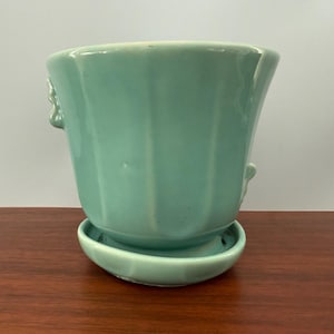May include: A vintage, light-green ceramic planter with a matching saucer. The planter has a fluted design and small, decorative handles. This piece is ideal for small plants and adds a touch of retro charm to any space.