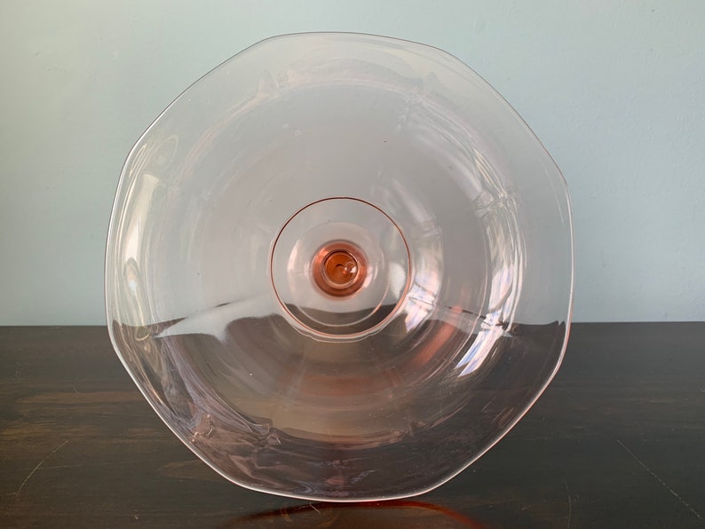May include: A clear glass cake stand with a pink tint and a scalloped edge. The stand has a raised center base.