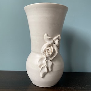 May include: White ceramic vase with a raised floral design. The vase has a wide mouth and a rounded body.