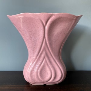 May include: A pink ceramic vase with a speckled finish and a unique heart-shaped design. The vase has a wide opening and a narrow base.