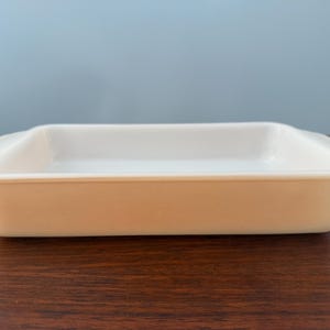 May include: A rectangular, off-white baking dish with rounded corners and handles. The dish is made of a solid material and sits on a dark brown wooden surface. The background is a solid light blue.