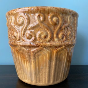 May include: A brown ceramic planter with a decorative embossed design. The planter has a wide, round opening and a slightly tapered body.