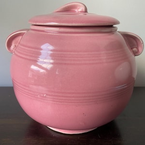 May include: A pink ceramic cookie jar with a lid and two handles. The jar has a smooth, glossy finish and is decorated with horizontal lines.