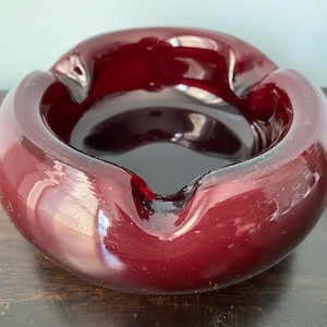 May include: A deep burgundy glass ashtray with a rounded, organic shape. The ashtray has a smooth, glossy finish.