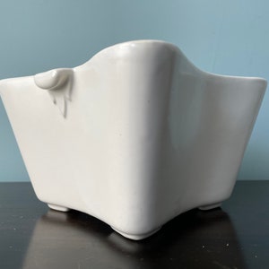 May include: White ceramic planter with a unique, wavy, star-shaped design. The planter has a small handle on one side.