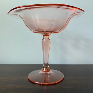 May include: A pink glass pedestal bowl with a scalloped edge and a fluted design. The bowl is on a wooden surface.