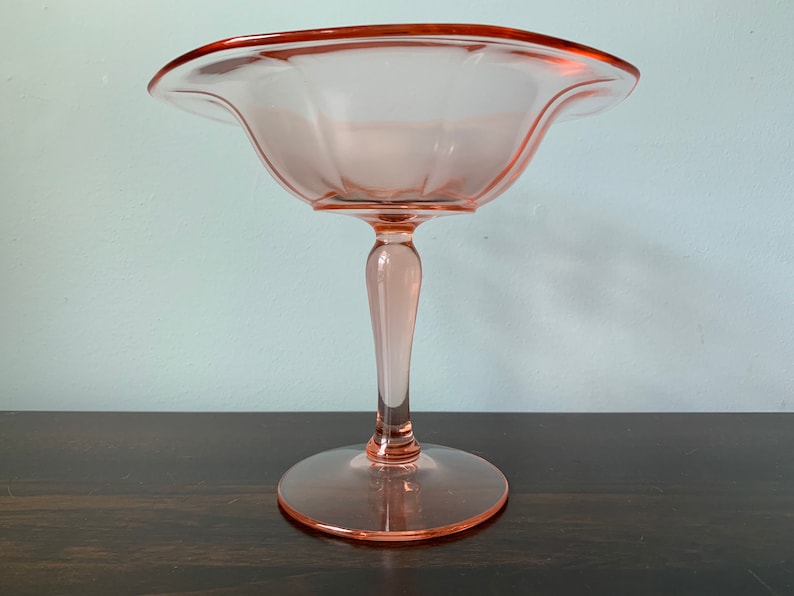 May include: A pink glass pedestal bowl with a scalloped edge and a fluted design. The bowl is on a wooden surface.
