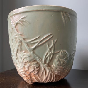 May include: A green ceramic planter with a raised design of pine cones and branches. The planter has a rounded shape and a slightly flared rim.
