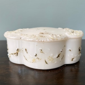 May include: White porcelain trinket box with a scalloped edge and gold floral designs. The box is closed.