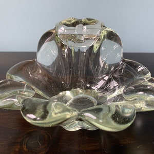 May include: A clear glass ashtray shaped like a flower with five petals. The ashtray has a raised center and a smooth, polished finish.
