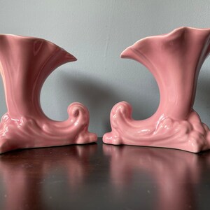 May include: Two pink ceramic vases with a trumpet-like opening and a decorative, wave-like base.
