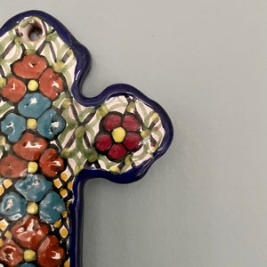 May include: A ceramic cross with a blue border and a floral pattern in shades of red, yellow, blue, and green.