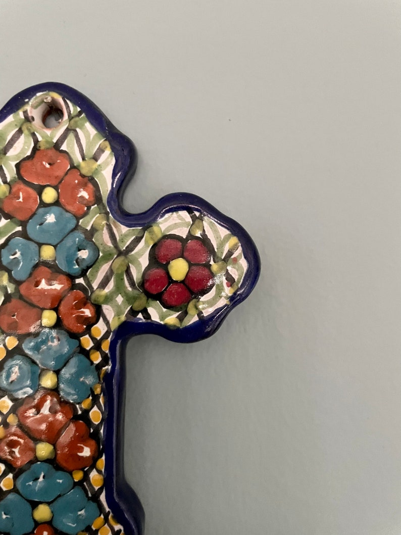 May include: A ceramic cross with a blue border and a floral pattern in shades of red, yellow, blue, and green.