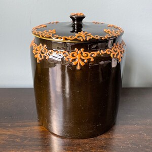 May include: A dark brown ceramic jar with a lid. The jar has a decorative orange floral design around the top and bottom.