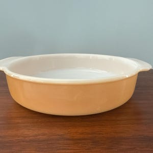 May include: An oval, light orange casserole dish with two small handles. The interior is a creamy white color. The dish is sitting on a dark brown wooden surface, with a light blue background.