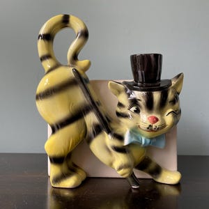 May include: A ceramic figurine of a cat wearing a top hat and a bow tie. The cat is yellow and black striped and has a playful expression. The figurine is holding a cane in its paw.