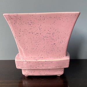 May include: A pink ceramic planter with a speckled finish. The planter has a square base and a wide, flared opening.