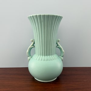 May include: A light green ceramic vase with a bulbous base and fluted, flared top. The vase has two ornate handles and measures approximately 10 inches tall.