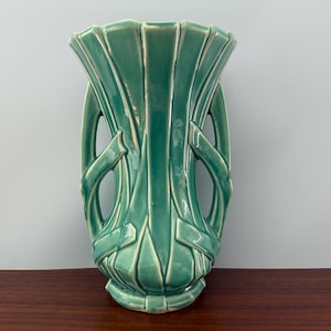 May include: A teal-colored ceramic vase with a fluted top and decorative cut-out handles. The vase has a textured surface and a pedestal base. The design is symmetrical and features vertical lines and geometric shapes. The vase is approximately 12 inches tall.