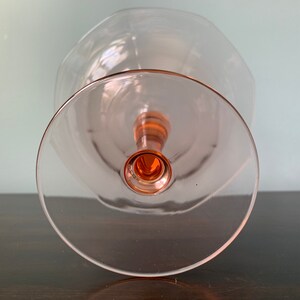 May include: A clear glass pedestal bowl with a pink rim and base. The bowl is empty and sits on a dark wooden surface.