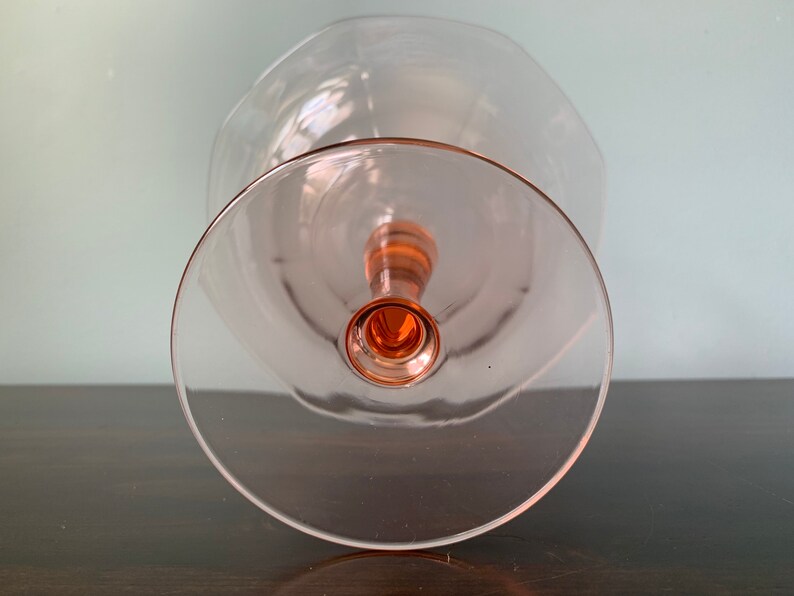 May include: A clear glass pedestal bowl with a pink rim and base. The bowl is empty and sits on a dark wooden surface.