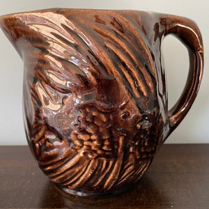 May include: A brown ceramic pitcher with a textured, swirling design. The pitcher has a wide mouth and a handle.