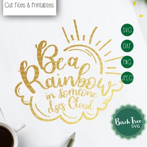 May include: Gold-colored text on white paper reads "Be a Rainbow in someone else's Cloud." The design includes a sun and cloud graphic. The image also shows file type icons: SVG, DXF, PNG, and JPEG.