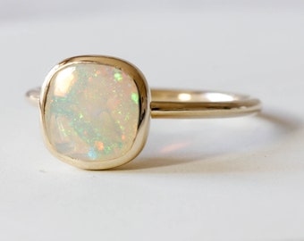 Può includere: Un anello d'oro con una pietra preziosa di opale bianco. L'opale ha una forma a cuscino ed è incastonata in una montatura a  bezel.