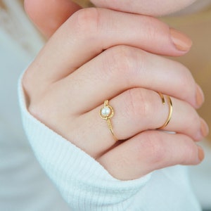 Puede incluir: Un anillo de oro con un pequeño acento de perla. El anillo se lleva en el dedo anular de una mano.