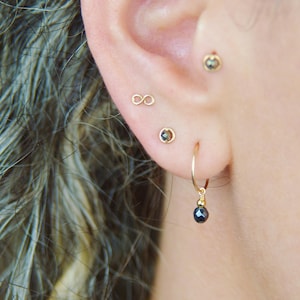 May include: A close-up of a person's ear with multiple piercings. The ear is pierced with a small infinity symbol, a small round gemstone, and a larger hoop earring with a dangling black gemstone.