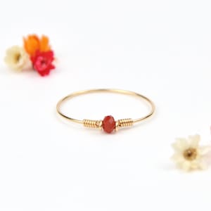 Op de afbeelding: Een delicate gouden ring met een kleine, gefacetteerde, rood-oranje edelsteen in het midden. De edelsteen is vastgezet met gouddraad. De ring staat tegen een witte achtergrond met kleurrijke bloemen.
