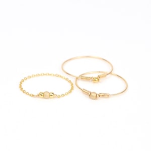 Op de afbeelding: Drie delicate gouden ringen worden getoond tegen een witte achtergrond. Eén ring is een ketting met een centrale kraal, de andere twee zijn eenvoudige banden met decoratieve draad- en kraalaccenten. De sieraden zijn elegant en minimalistisch.